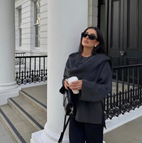 Frau in grauem Mantel mit Sonnenbrille und Kaffeebecher vor weißem Gebäude.