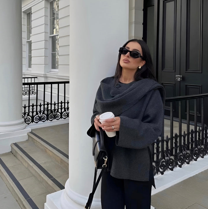 Frau in grauem Mantel mit Sonnenbrille und Kaffeebecher vor weißem Gebäude.