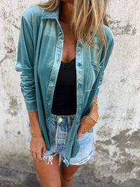 Frau in türkisfarbener Bluse, schwarzem Top und Jeansshorts vor Betonwand.