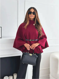 Frau in rotem Cape, schwarze Hose, Sonnenbrille, hält schwarze Handtasche, eleganter Look.