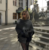 Frau in grauem Poncho und Sonnenbrille, schwarze Handschuhe, Tasche, städtische Kulisse.