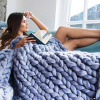 Frau mit Buch unter blauem, grob gestricktem Wollplaid auf Sofa, gemütliches Wohnzimmer.
