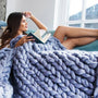 Frau mit Buch unter blauem, grob gestricktem Wollplaid auf Sofa, gemütliches Wohnzimmer.