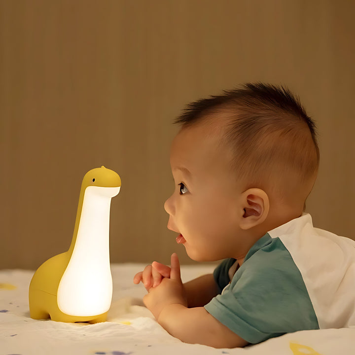 Gelbe Dinosaurier-Nachtlampe aus Silikon mit LED, neben Baby auf Bett, Kinderzimmer-Dekor.