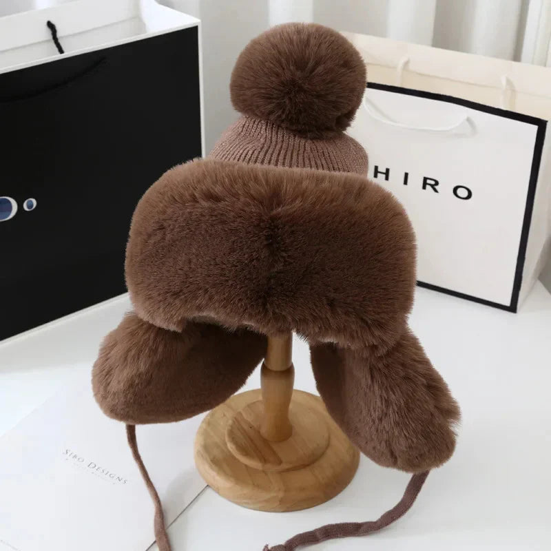 Braune Wintermütze mit Kunstfell, Bommel und Ohrenklappen auf Holzständer, modisch und warm.