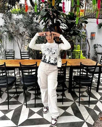 Frau in weißem Langarmshirt und Cargo-Hose posiert vor Holztisch im Garten mit Pflanzen.