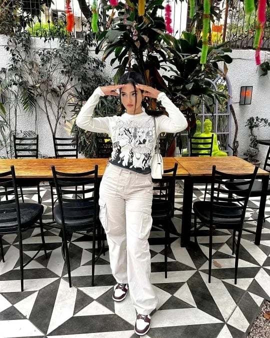 Frau in weißem Langarmshirt und Cargo-Hose posiert vor Holztisch im Garten mit Pflanzen.