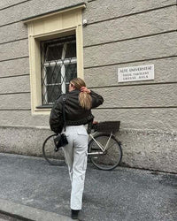 Frau in schwarzer Jacke und weißer Hose vor Universität, Fahrrad im Hintergrund.