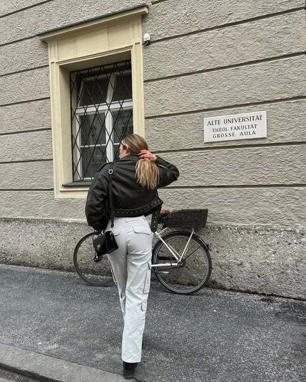 Frau in schwarzer Jacke und weißer Hose vor Universität, Fahrrad im Hintergrund.