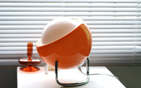 Orange Retro-Tischlampe mit weißem Globus, modernes Design, vor Jalousien auf Tisch.