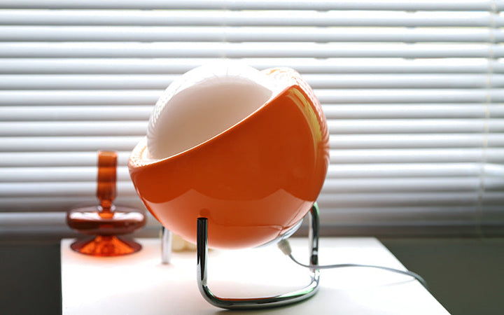 Orange Retro-Tischlampe mit weißem Globus, modernes Design, vor Jalousien auf Tisch.