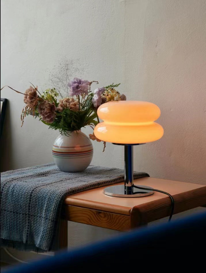 Moderne Tischlampe aus Glas, orange leuchtend, auf Holztisch neben Blumenstrauß in Vase.