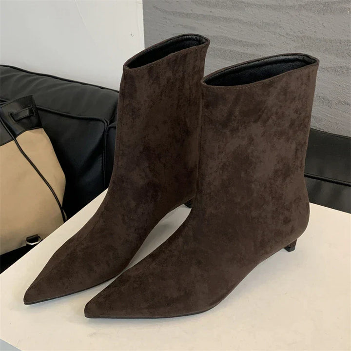 Dunkelbraune Wildleder-Stiefeletten mit spitzem Absatz auf weißem Tisch, modisch und elegant.