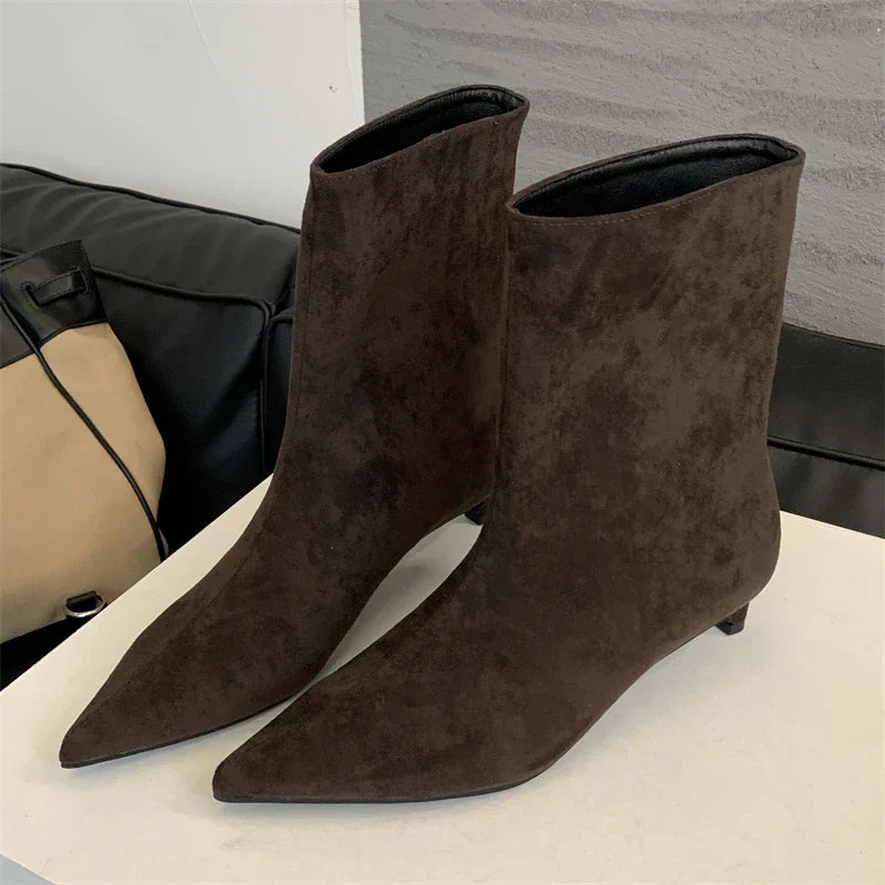 Dunkelbraune Wildleder-Stiefeletten mit spitzem Absatz auf weißem Tisch, modisch und elegant.
