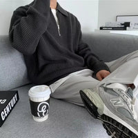 Mann in grauem Strickpullover mit Reißverschluss, weiße Hose, graue Sneaker, Kaffeebecher.
