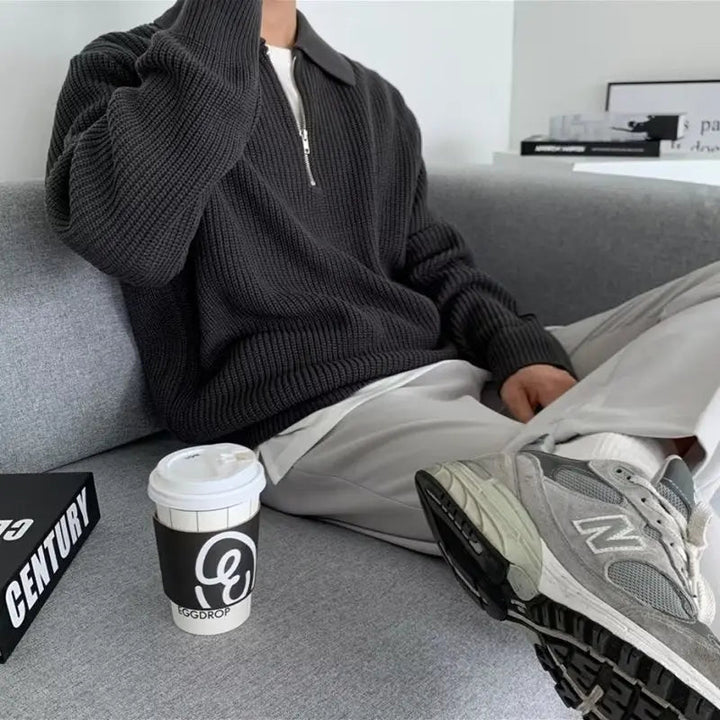 Mann in grauem Strickpullover mit Reißverschluss, weiße Hose, graue Sneaker, Kaffeebecher.