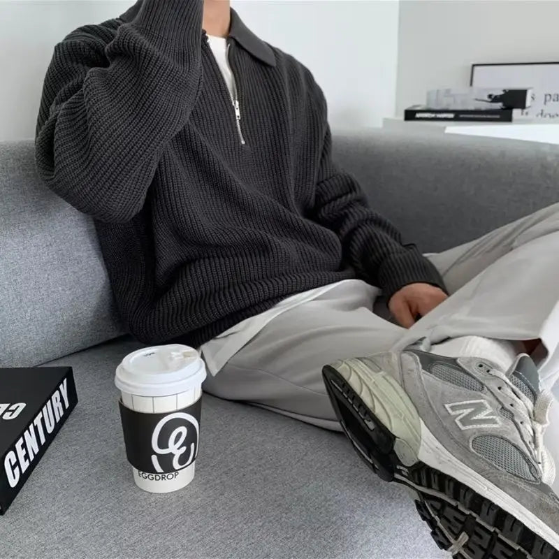 Mann in grauem Strickpullover mit Reißverschluss, weiße Hose, graue Sneaker, Kaffeebecher.
