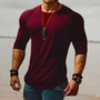 Mann in bordeauxrotem Langarmshirt, muskulös, mit Halskette und Armbändern am Strand.