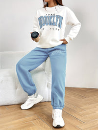 Frau in weißem "Brooklyn" Sweatshirt und blauer Jogginghose, hält schwarzen Kaffeebecher.