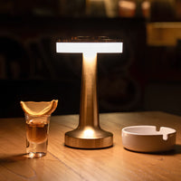 Goldene Tischlampe mit modernem Design neben einem Glas mit Orangenscheibe und weißem Aschenbecher.