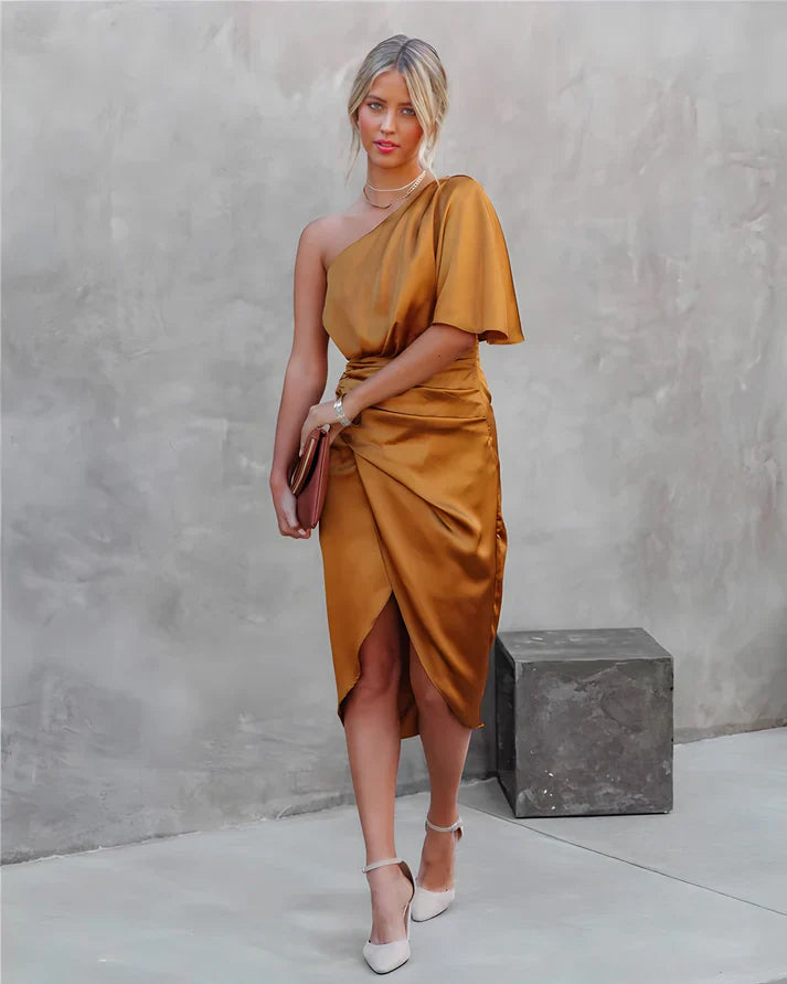 Frau in einem goldenen, schulterfreien Satin-Kleid mit Clutch und weißen High Heels vor grauer Wand.