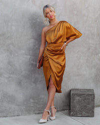 Frau in goldfarbenem Satin-Kleid, asymmetrisch, mit Clutch und High Heels vor Betonwand.