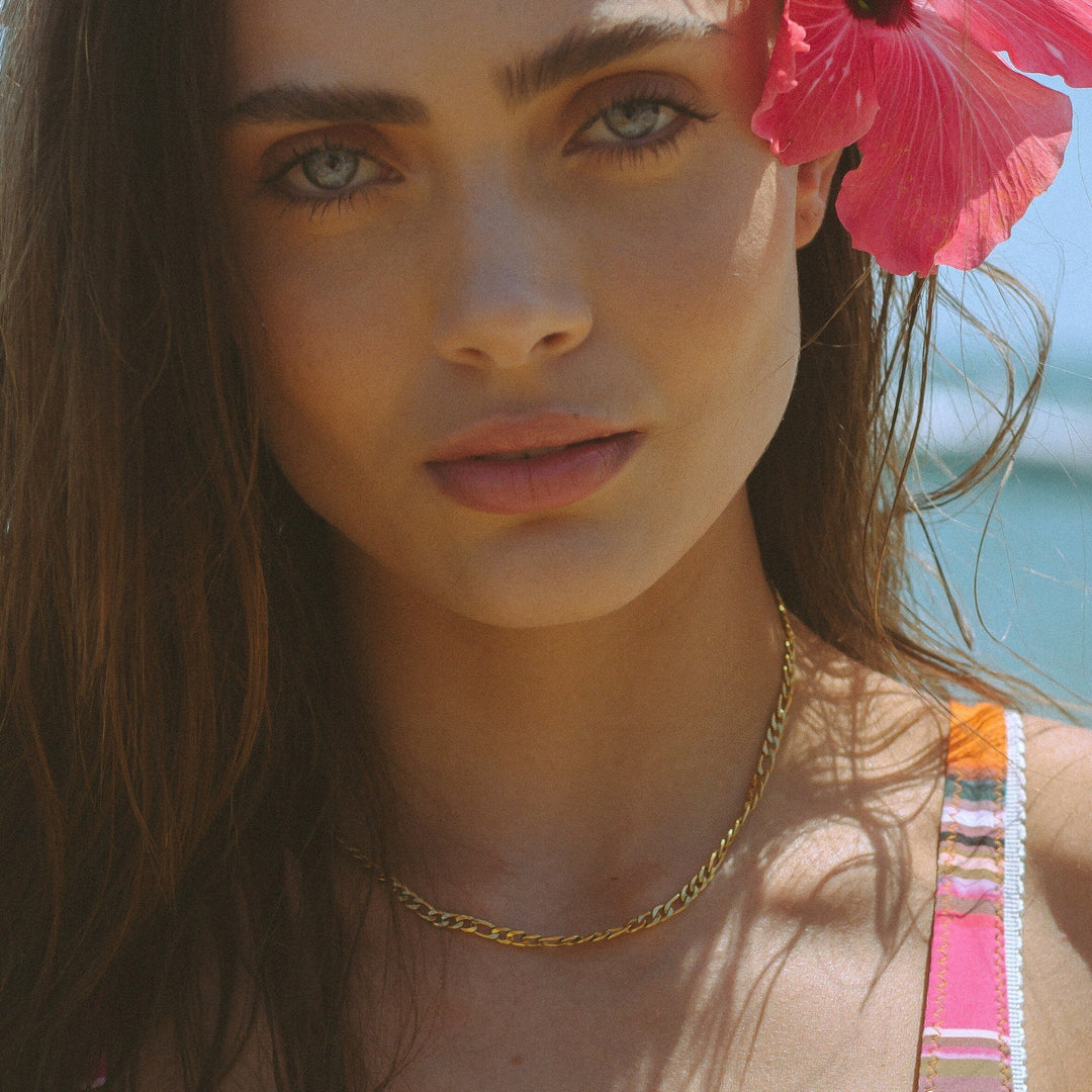 Frau mit goldener Figaro-Halskette und roter Hibiskusblüte im Haar, Sommermode.