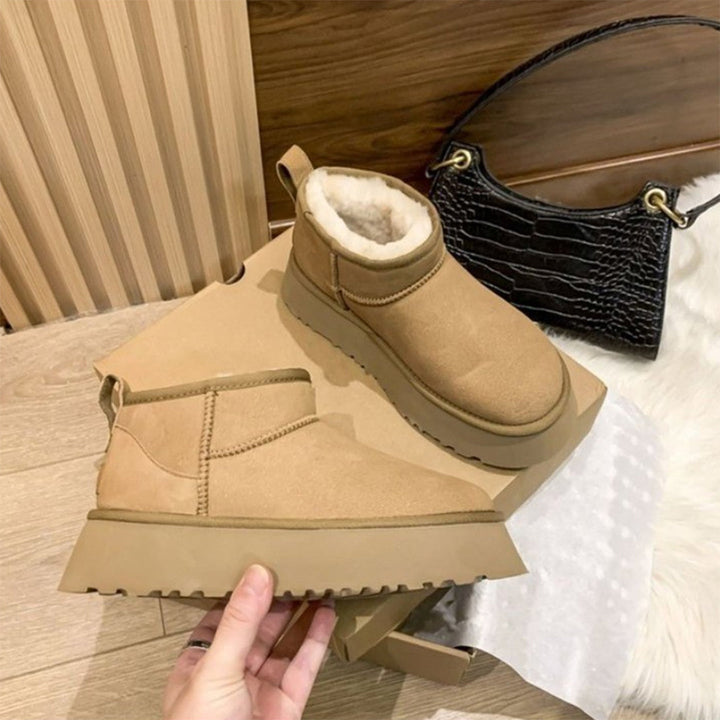 Beige Winterstiefel aus Wildleder mit Fellfutter auf Karton, schwarze Handtasche im Hintergrund.