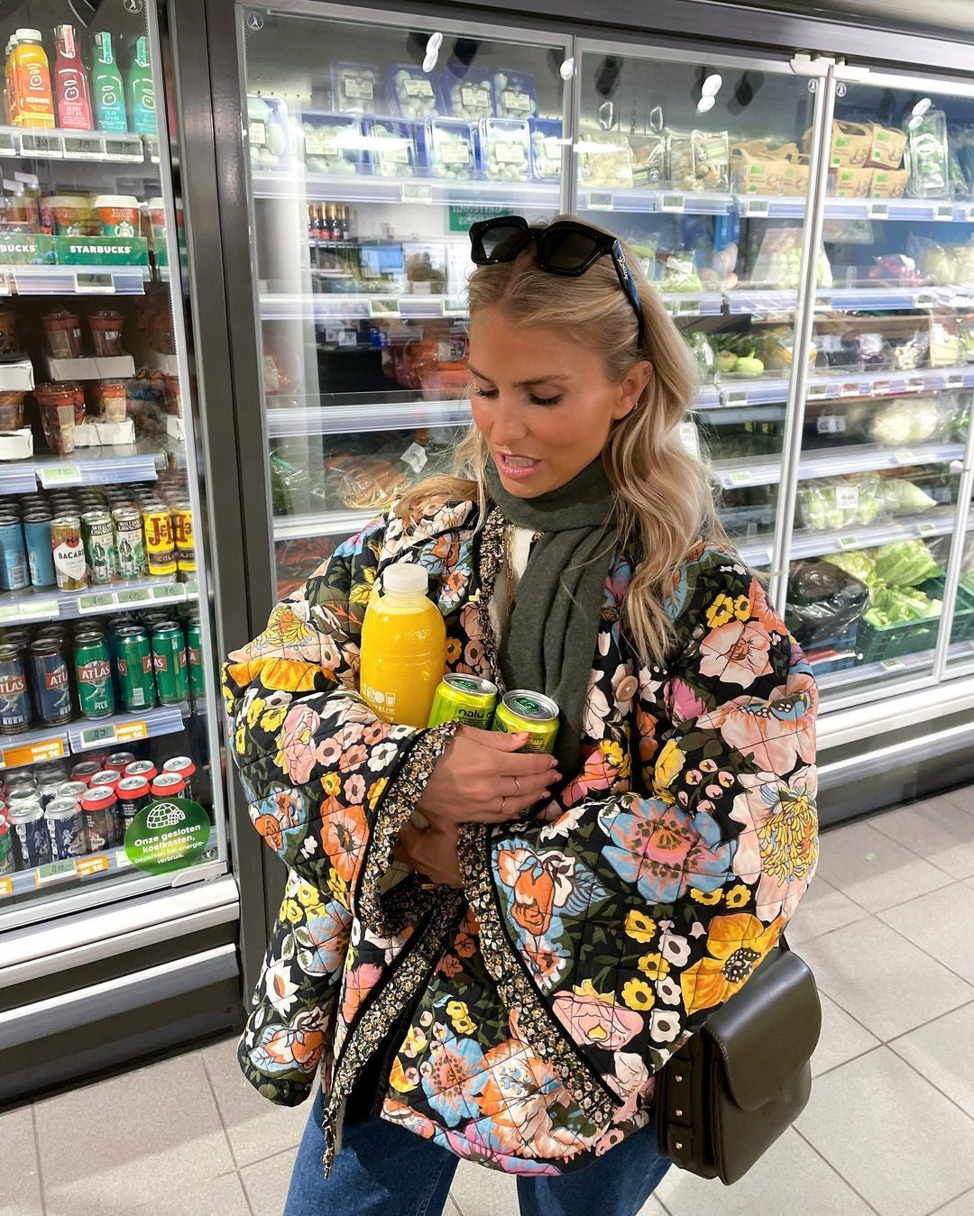 Frau im Supermarkt mit bunter Blumenjacke hält Orangensaft und Getränke vor Kühlregal.
