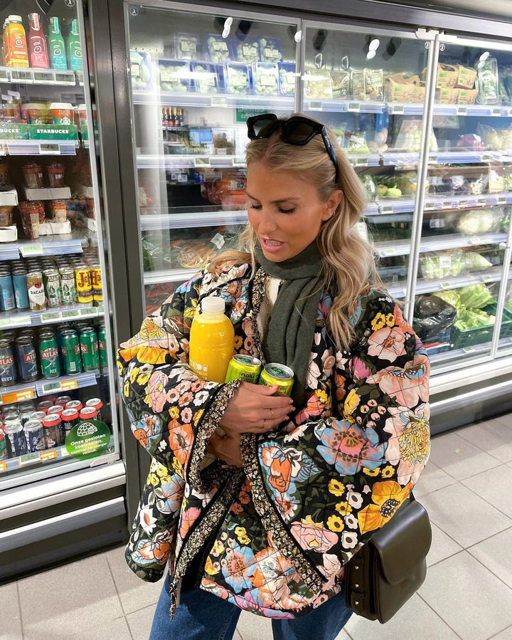 Frau im Supermarkt mit bunter Blumenjacke, hält Orangensaft und Dosen, vor Kühlregal.