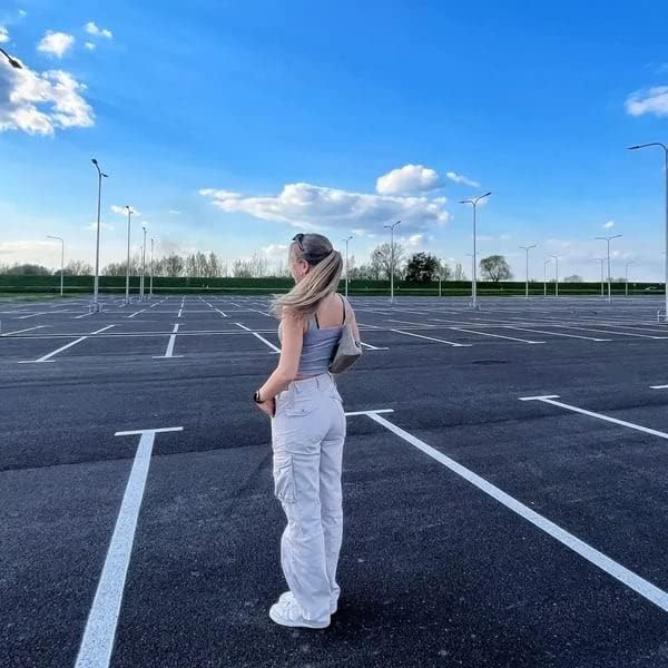 Frau in weißer Hose und grauem Top steht auf leerem Parkplatz unter blauem Himmel.