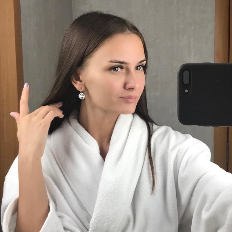 Frau mit weißen Ohrringen, Selfie im Spiegel, weißer Bademantel, lange braune Haare.