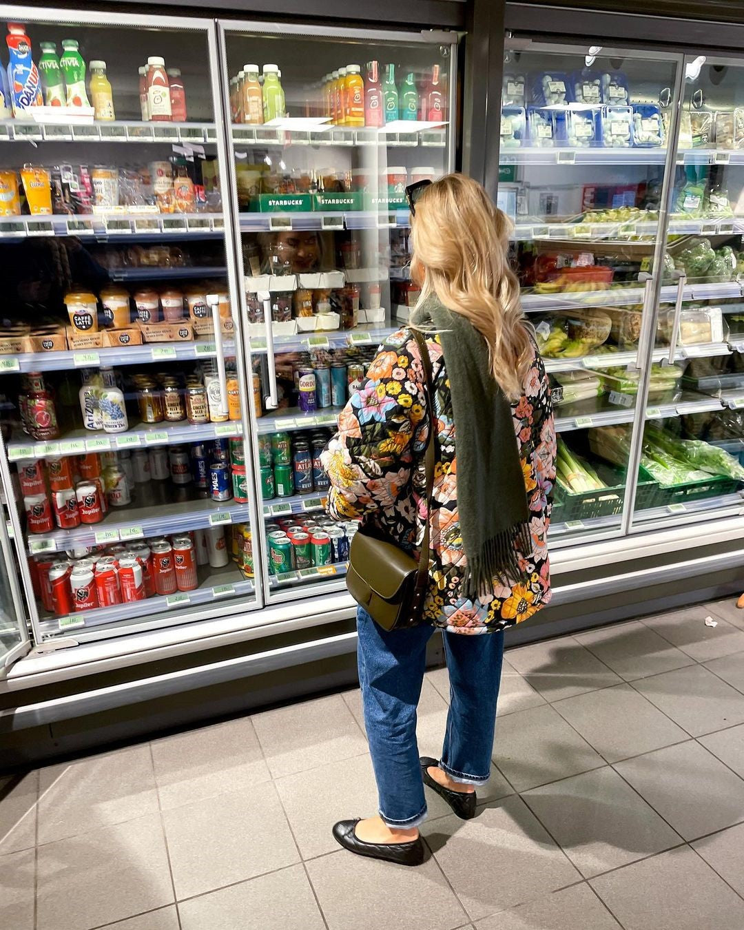 Frau im Supermarkt vor Kühlregal mit Getränken und Lebensmitteln, Blumenjacke, Jeans, Schal.