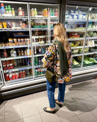 Frau im Supermarkt vor Kühlregal mit Getränken und Lebensmitteln, Blumenjacke, Jeans, Schal.
