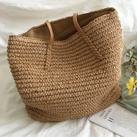 Große beige Strohtasche mit Ledergriffen, ideal für Strand oder Einkauf, neben Blumen und Buch.