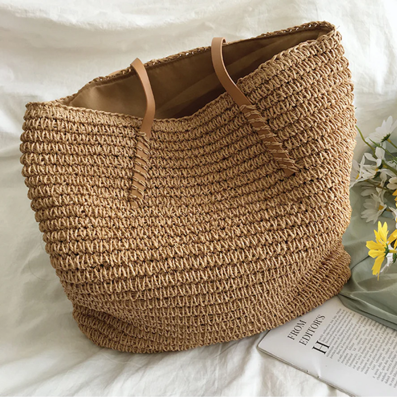 Große beige Strohtasche mit Ledergriffen, ideal für Strand oder Einkauf, neben Blumen und Buch.