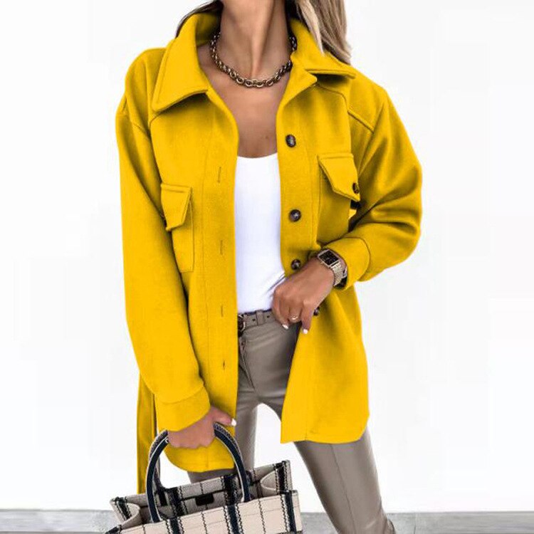 Frau in gelber Jacke, weiße Bluse, beige Hose, hält karierte Handtasche.