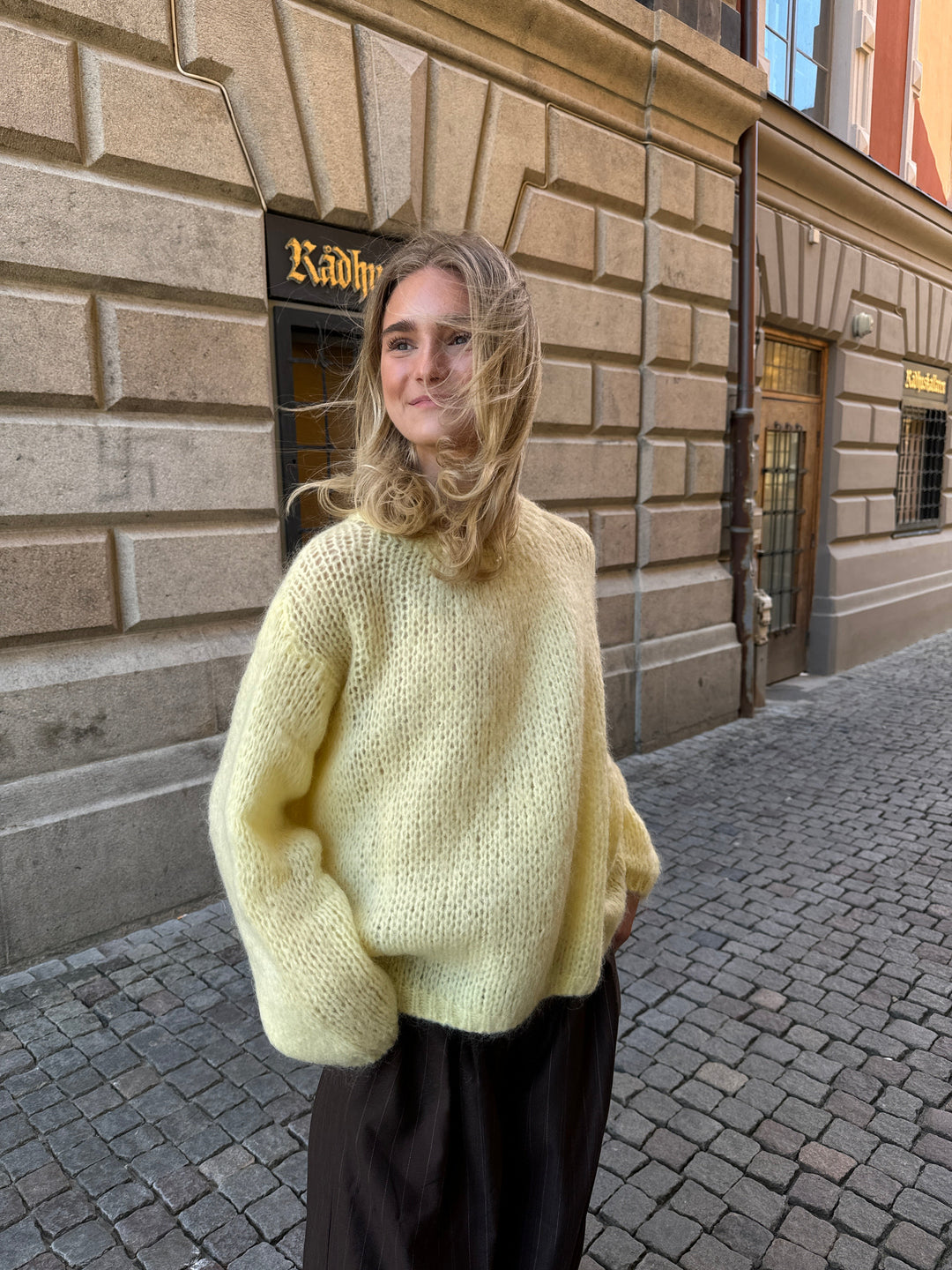 Frau in gelbem Strickpullover auf Kopfsteinpflasterstraße vor historischer Fassade.