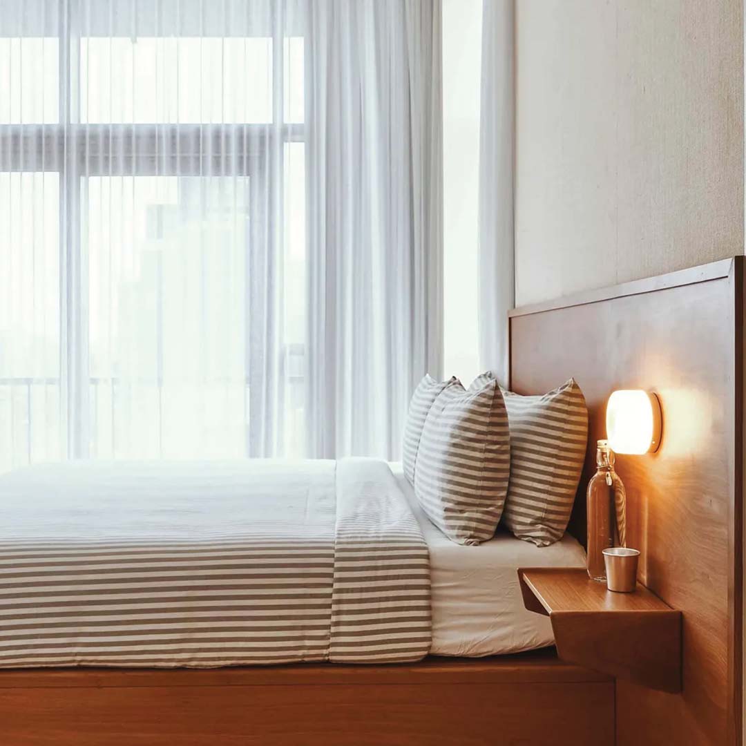 Modernes Schlafzimmer mit Holz-Doppelbett, gestreifter Bettwäsche, Nachttischlampe, Tageslicht.