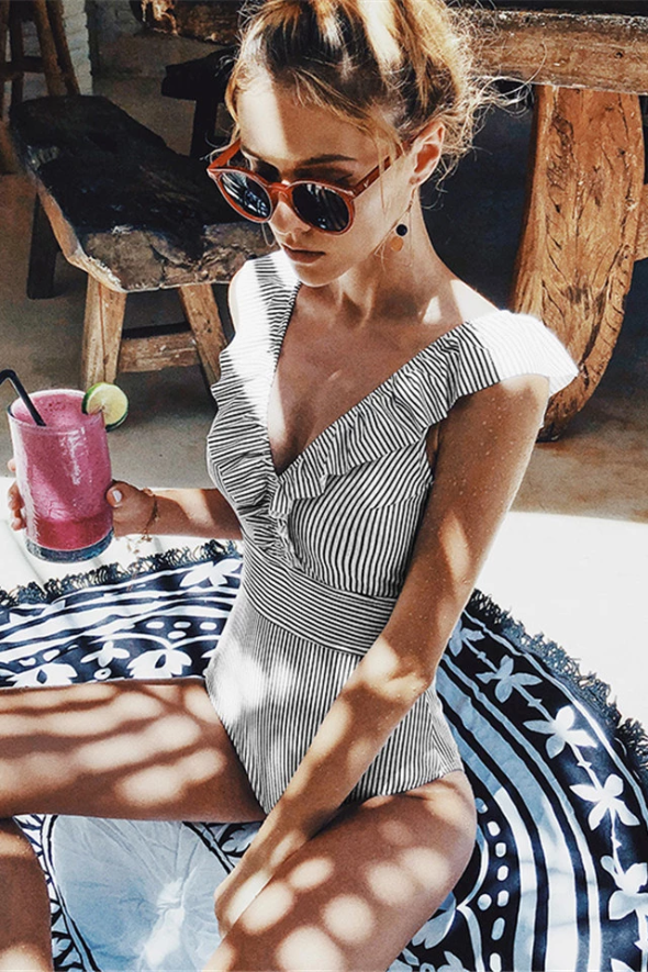 Frau im gestreiften Badeanzug mit Sonnenbrille, hält pinken Smoothie, sitzt auf gemustertem Tuch.