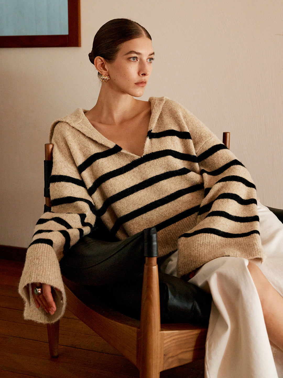 Frau in gestreiftem Pullover, beige mit schwarzen Streifen, sitzt auf Stuhl, modisch, elegant.