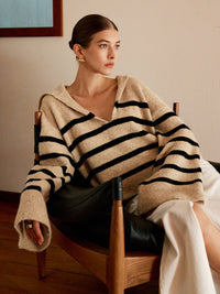 Frau in gestreiftem Pullover, beige mit schwarzen Streifen, sitzt auf Stuhl, modisch, elegant.