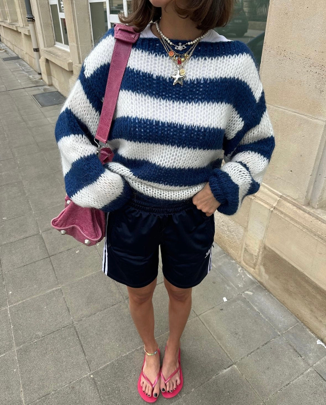 Frau in gestreiftem Pullover, blauer Shorts, rosa Tasche und Flip-Flops auf Gehweg.