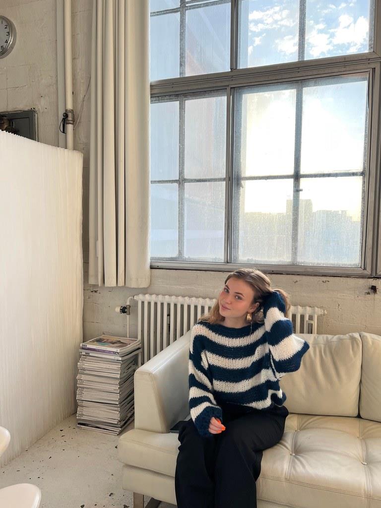 Frau in gestreiftem Pullover sitzt auf weißem Sofa vor großem Fenster in modernem Raum.