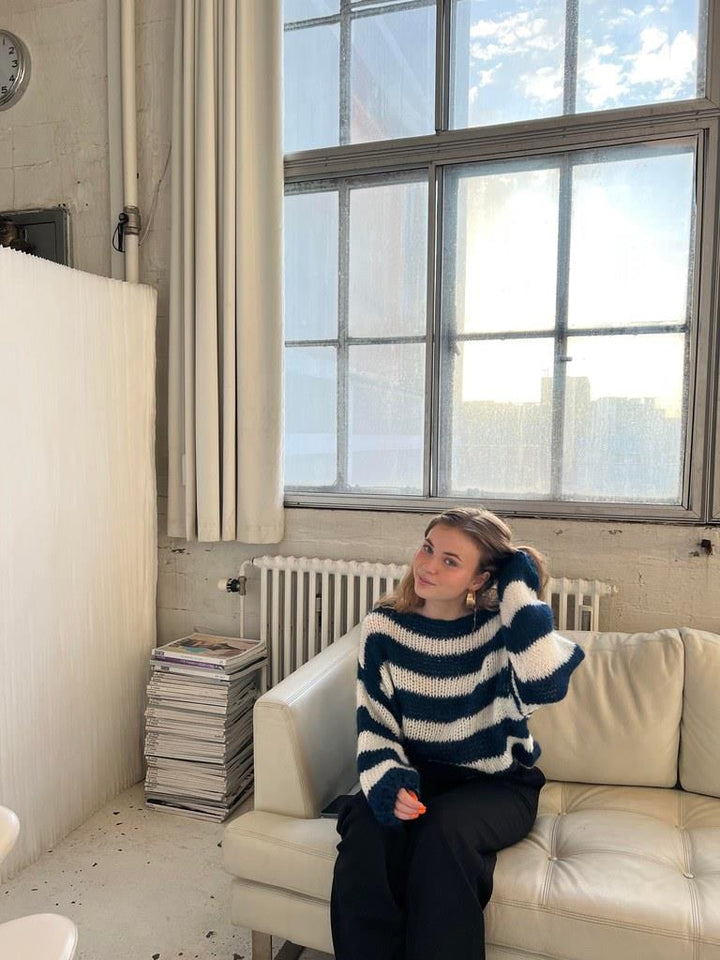 Frau in gestreiftem Pullover sitzt auf weißem Sofa vor großem Fenster in modernem Raum.