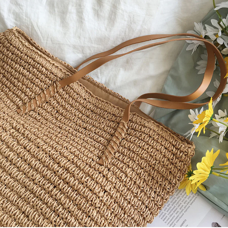 Geflochtene Strohtasche mit braunen Ledergriffen, ideal für Sommer und Strand, auf Blumenhintergrund.