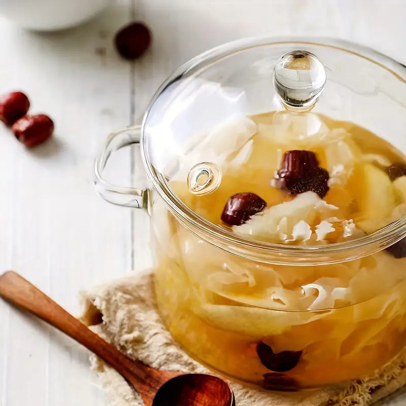 Glas-Teekanne mit Deckel, gefüllt mit Kräutertee und roten Datteln auf weißem Holztisch.