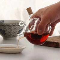 Hand gießt Tee aus Glas-Teekanne mit Korken in Schale, traditionelles Teeservice, transparent.