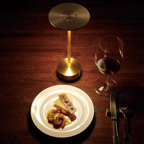 Goldene Tischlampe aus Metall auf Holztisch neben Teller mit Essen und Glas Rotwein.