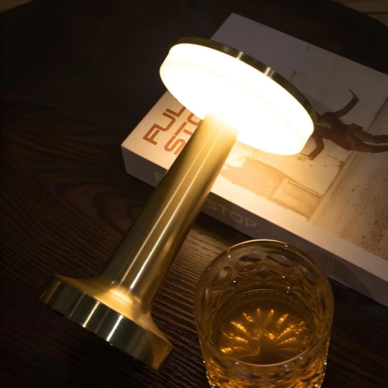 Goldene Tischlampe aus Metall, leuchtend, neben Kristallglas und Buch auf dunklem Holztisch.